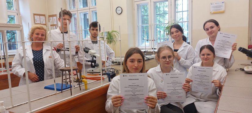 Uczestnicy wraz z opiekunką w laboratorium pozują do zdjęcia z dyplomami trzymanymi w rękach.