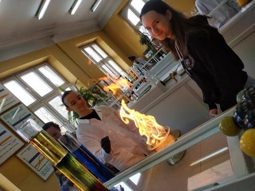 Uczniowie przeprowadzają doświadczenie dla gości w laboratorium chemicznym. Wjego wyniku widać duży płomień na pierwzym planie. PS. Nikomu nie stała się krzywda.